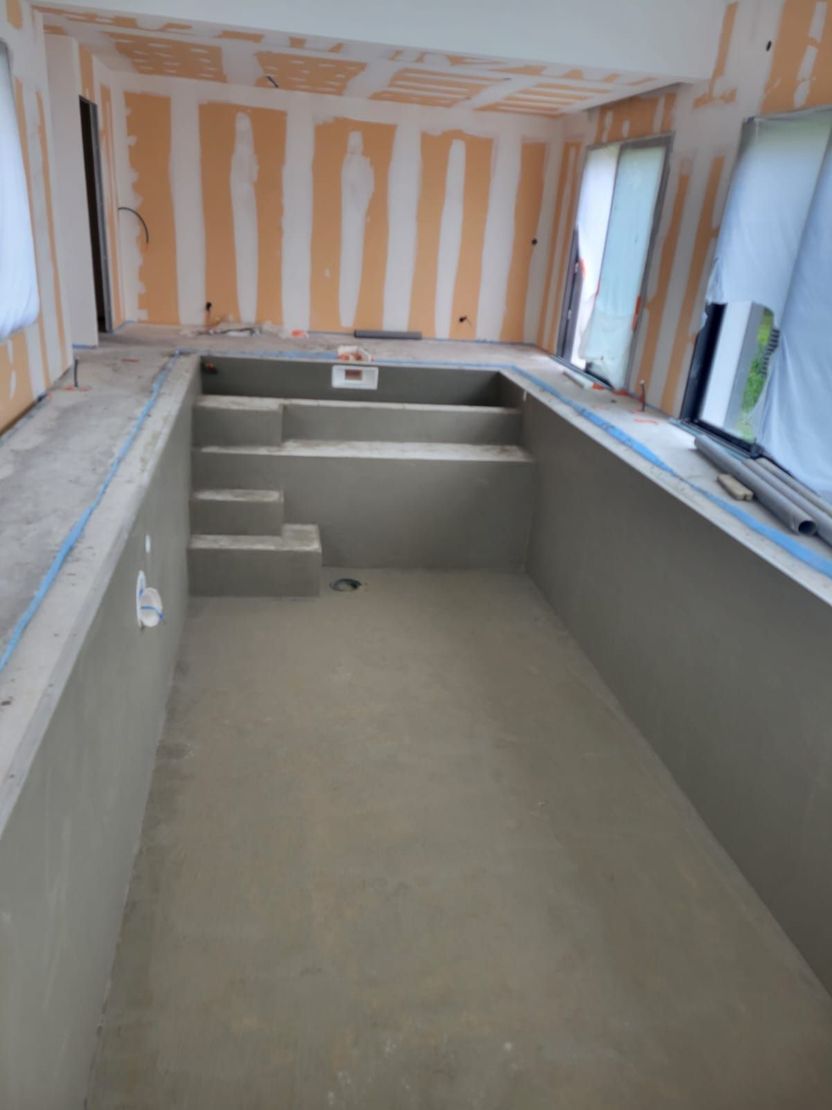 Piscine partiellement construite dans une cour arrière, avec murs en béton, jouxtant une maison beige et un muret paysager en pierre.
