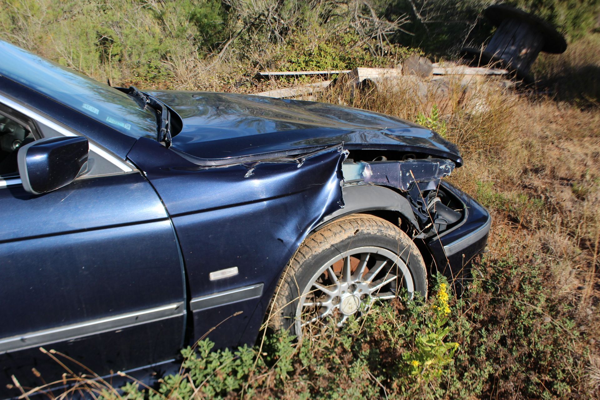 Voiture bleue avec l'avant endommagé, hors de la route, dans des herbes sèches et des broussailles.