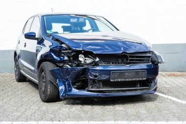 Une berline bleue endommagée, présentant des dégâts à l'avant gauche suite à une collision, est stationnée dans la rue.