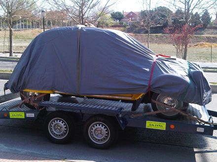Automóvil cubierto con una lona sobre un remolque, probablemente transportado. Automóvil amarillo, lona gris, remolque azul.