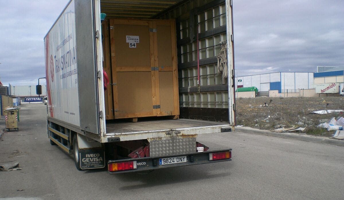 Un camión con la parte trasera abierta, con una gran caja en su interior, estacionado en una carretera con edificios industriales al fondo.