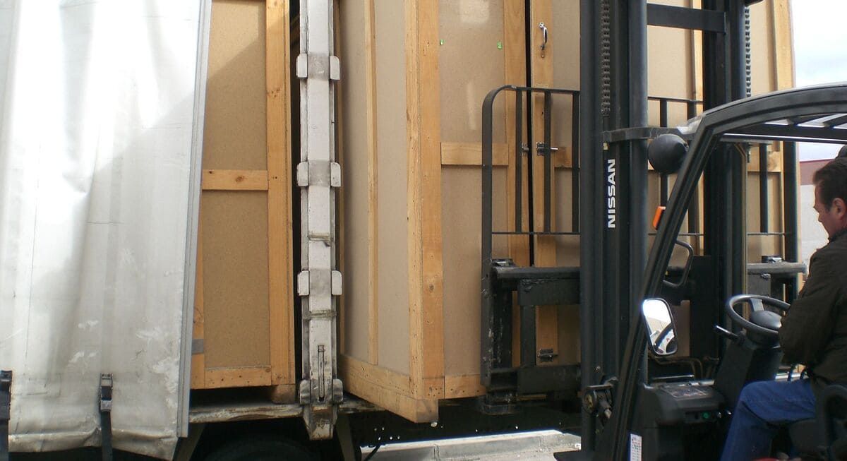 Una carretilla elevadora carga un objeto grande con estructura de madera en un camión. Una persona la opera.