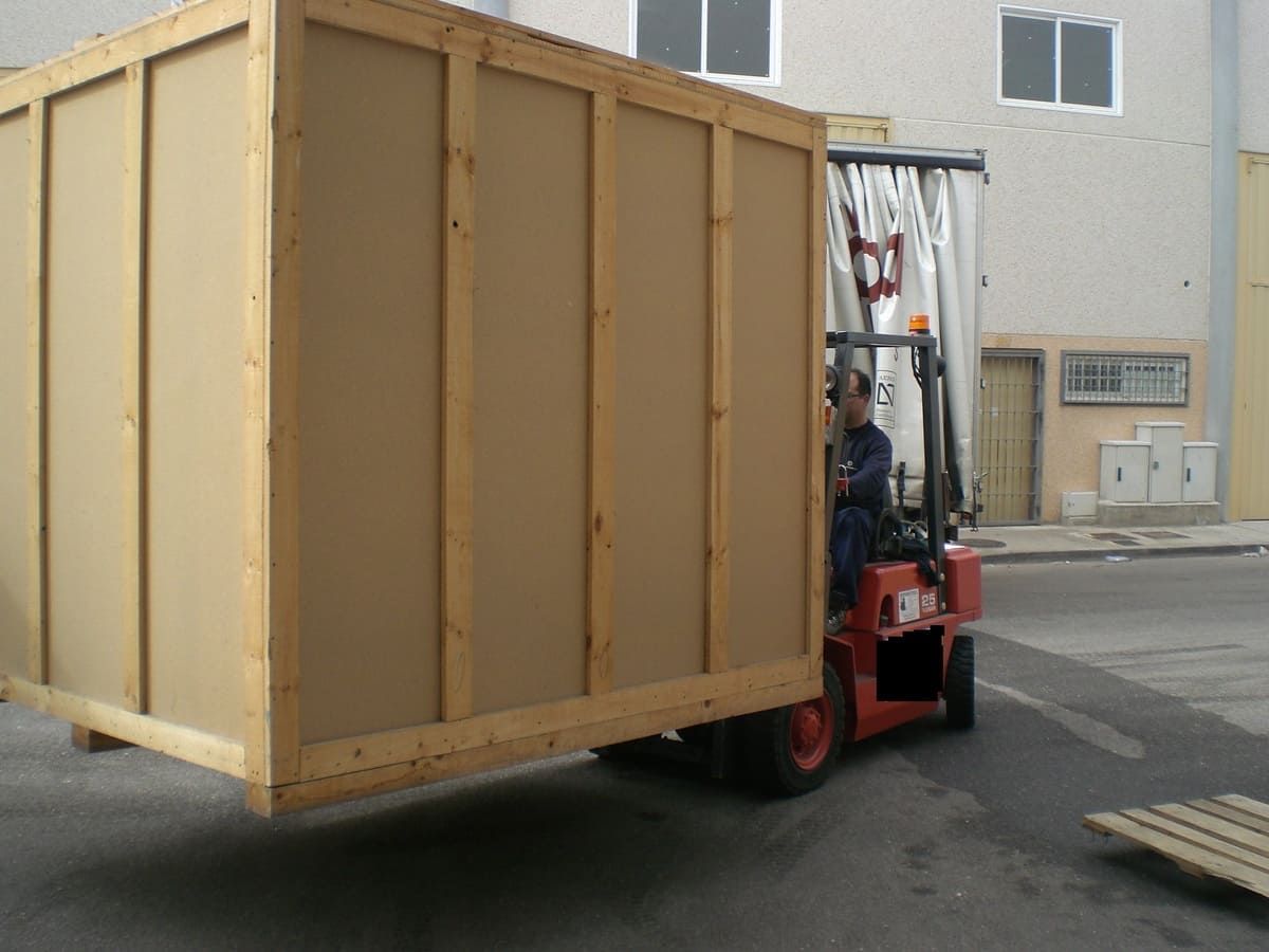 Carretilla elevadora que transporta una gran caja de madera al aire libre, con un trabajador conduciendo.