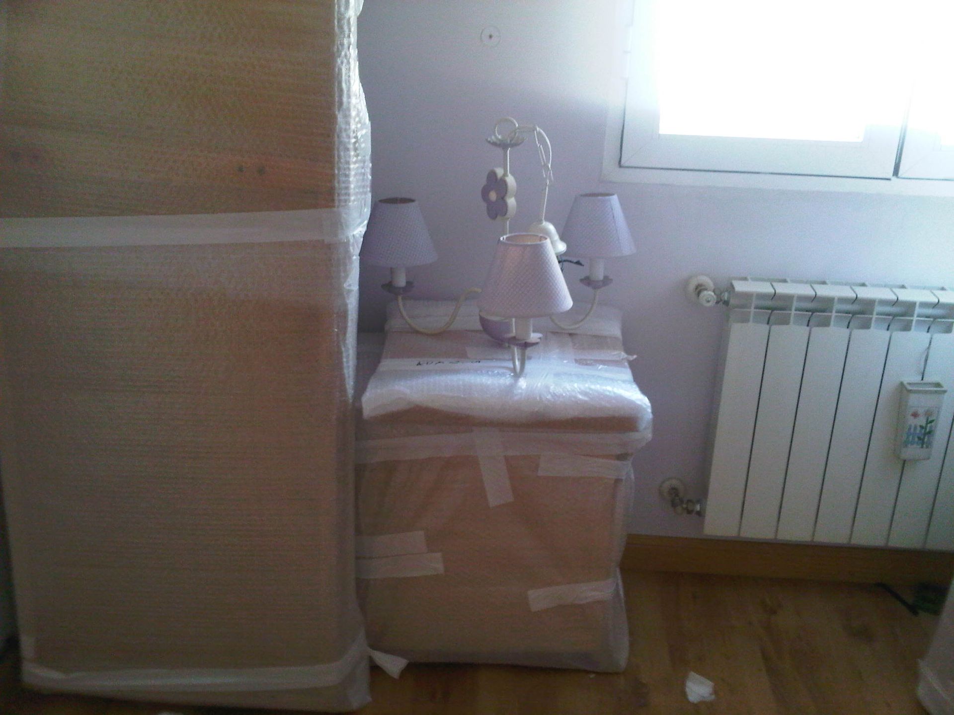 Unas cajas y una mesita de noche envueltas se encuentran en una habitación con radiador y ventana.