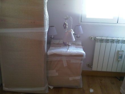 Unas cajas y una mesita de noche envueltas se encuentran en una habitación con radiador y ventana.