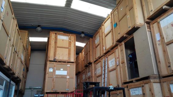 Interior de almacén lleno de cajas de madera apiladas. Luces fluorescentes, techo metálico y luz natural procedente de claraboyas.
