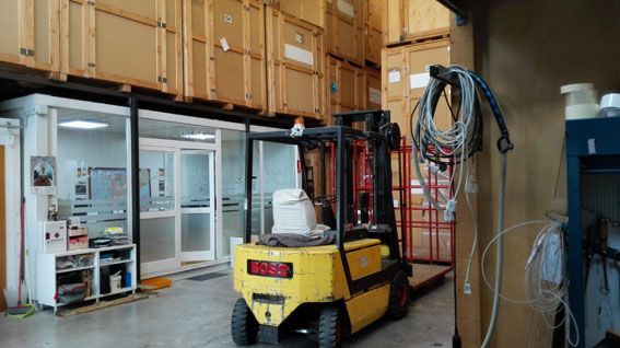 Una carretilla elevadora amarilla en un almacén, con cajas apiladas. Se ve una puerta abierta y cables eléctricos colgados de la pared.