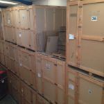 Filas de grandes cajas de madera apiladas en un almacén, algunas con etiquetas, posiblemente para almacenamiento o envío.