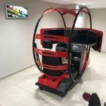 Simulador de conducción de realidad virtual rojo y negro en una habitación blanca con un televisor en la pared.