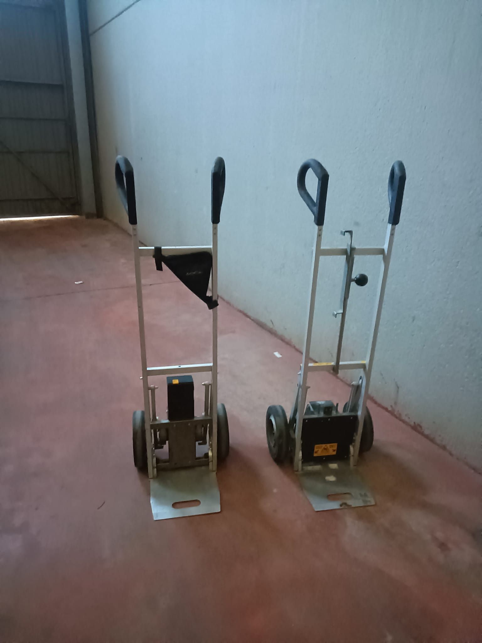 Dos carretillas manuales de aluminio con manijas y ruedas negras sobre un piso de concreto rojo.