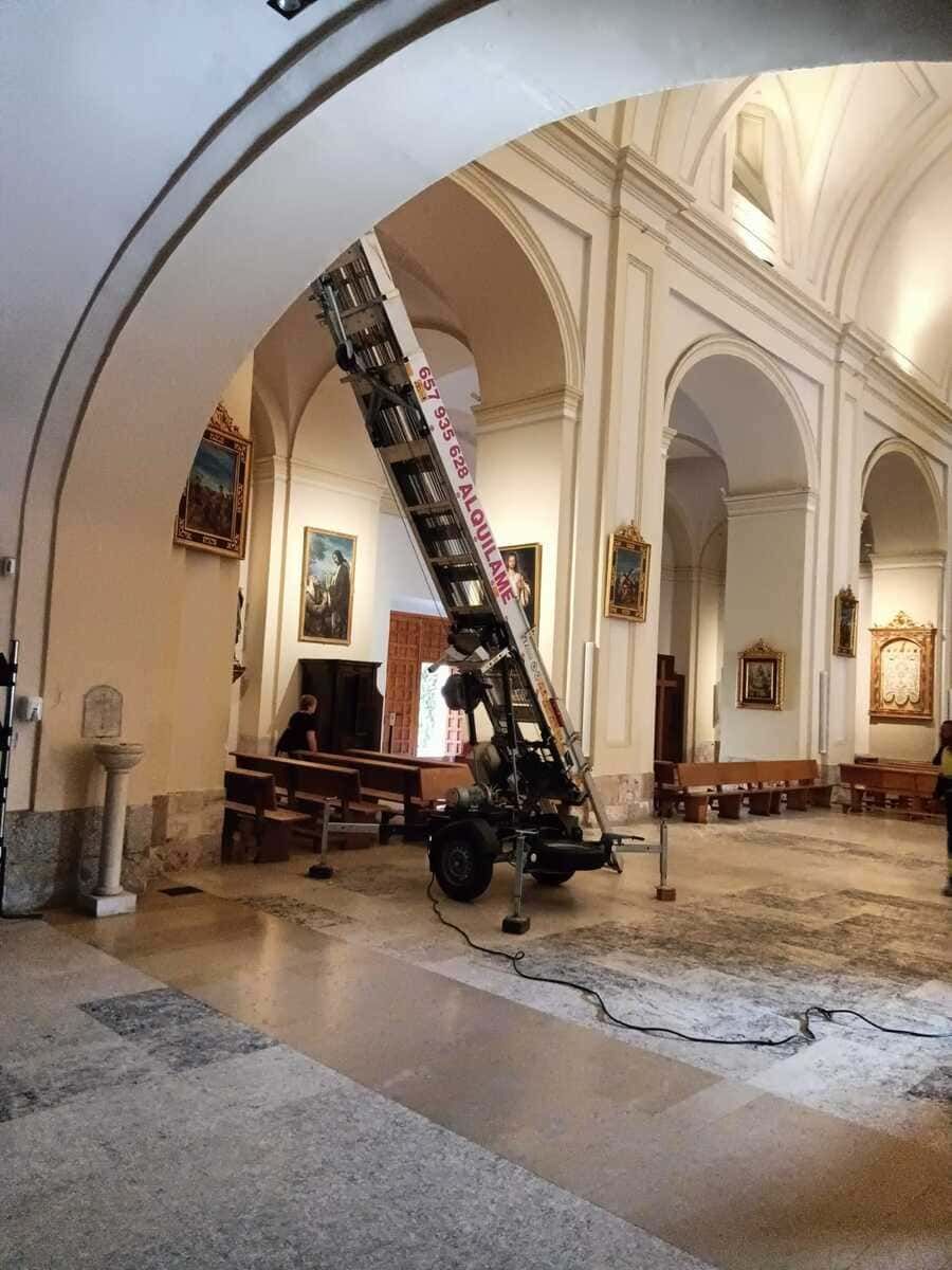 Un ascensor industrial dentro de una iglesia. Se ven arcos blancos, pinturas y bancos.