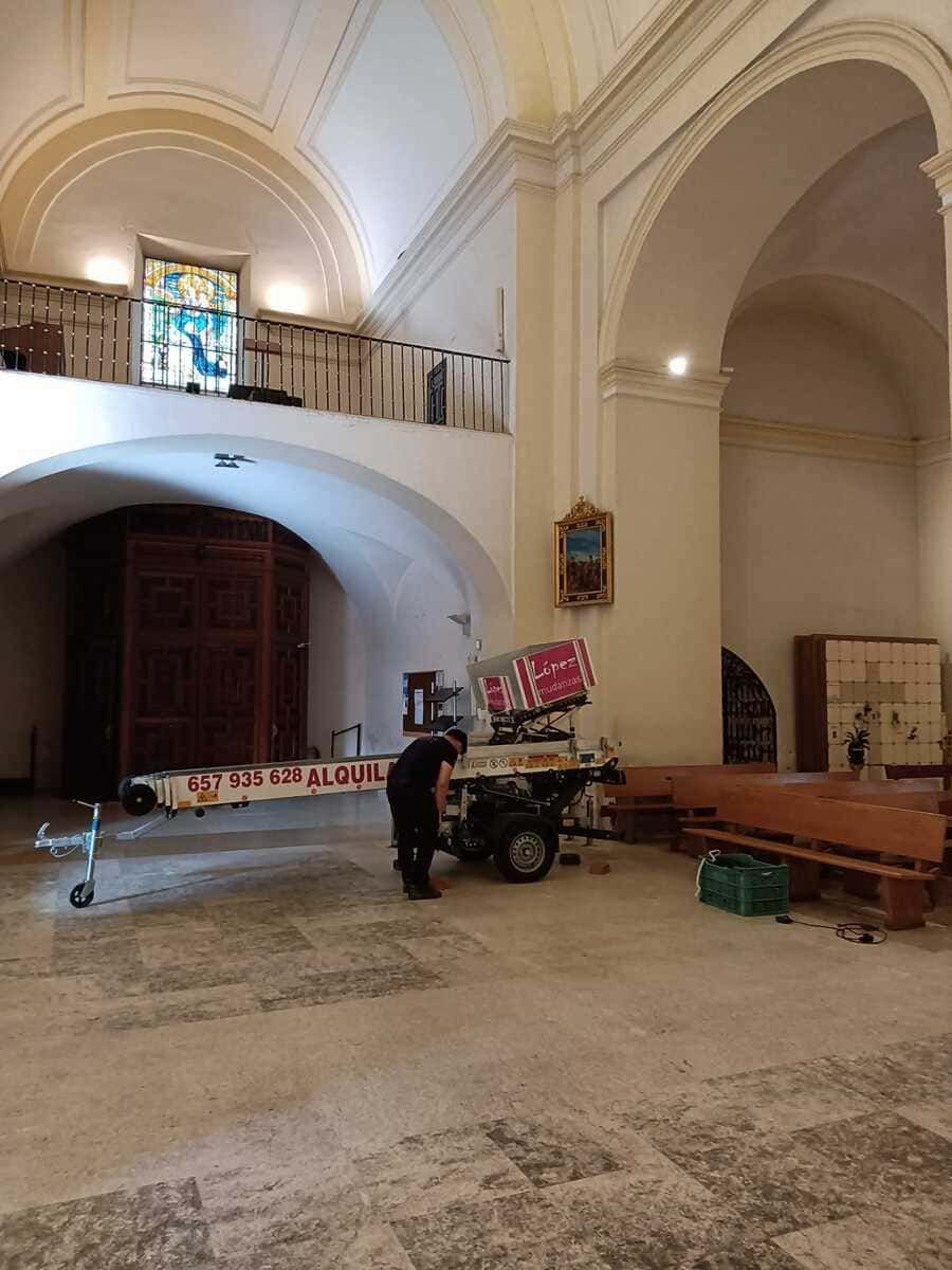 Dentro de una iglesia, una persona ajusta un ascensor. Bancos de madera bordean la pared, la puerta arqueada y el balcón.