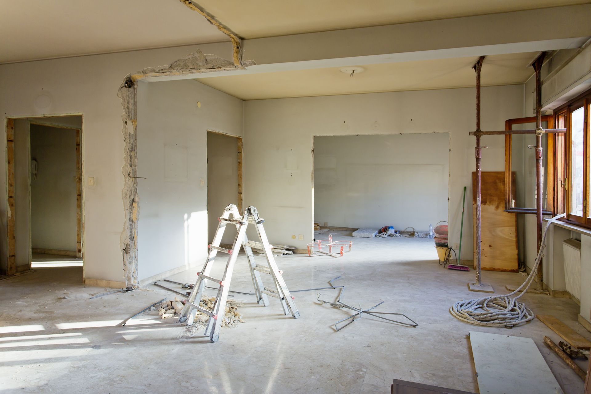 Pièce intérieure en cours de rénovation ; murs apparents, échelle, débris, poutres de soutien et fenêtre.
