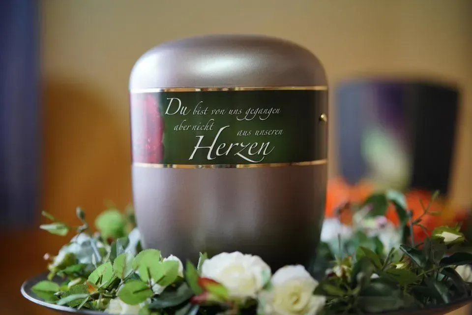 Eine silberne Urne mit einem Gedenkspruch ist von Blumen umgeben.