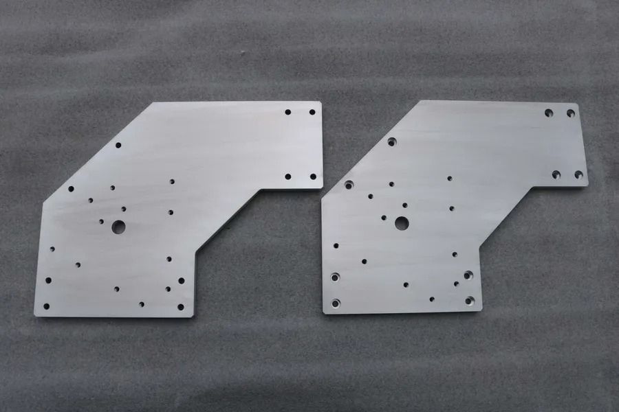 Zwei silberne Metallteile mit Bohrungen, nebeneinander auf einer grauen Oberfläche