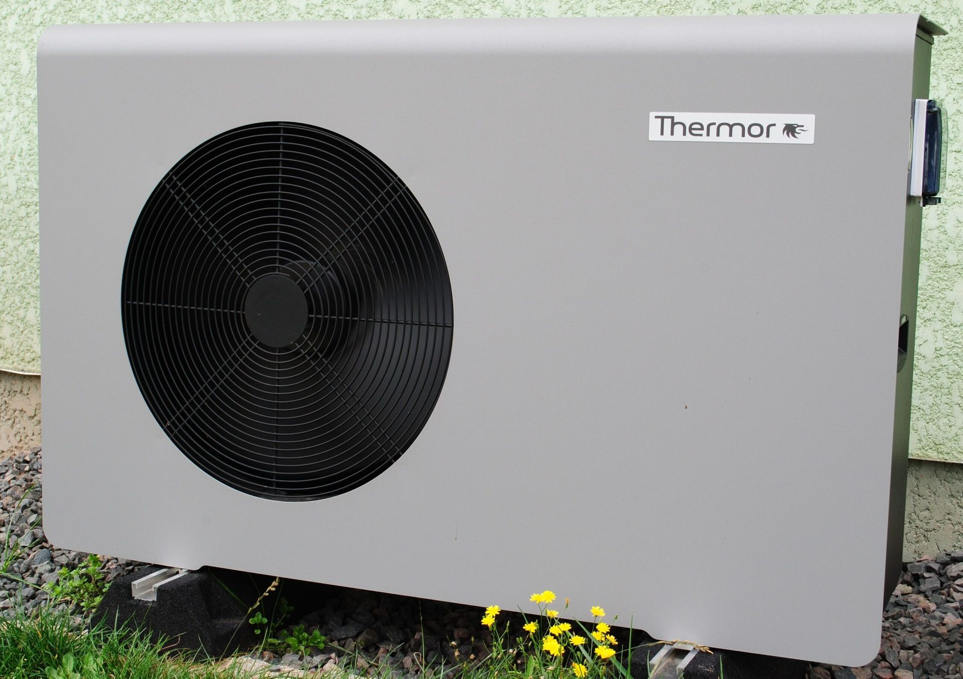 Unité de pompe à chaleur grise, ventilateur rond, montée sur blocs, à l'extérieur.