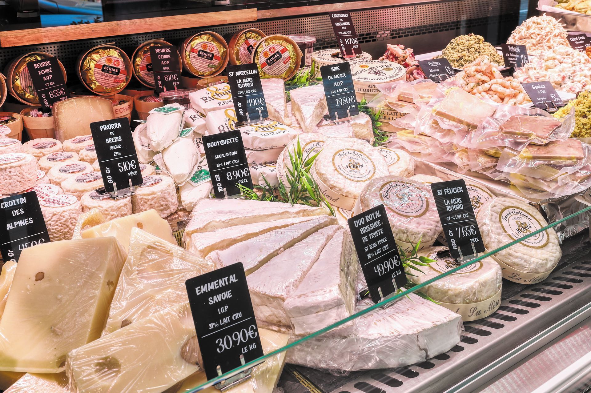 Comptoir de charcuterie avec assortiment de fromages, plateaux garnis et étiquettes de prix derrière une vitrine