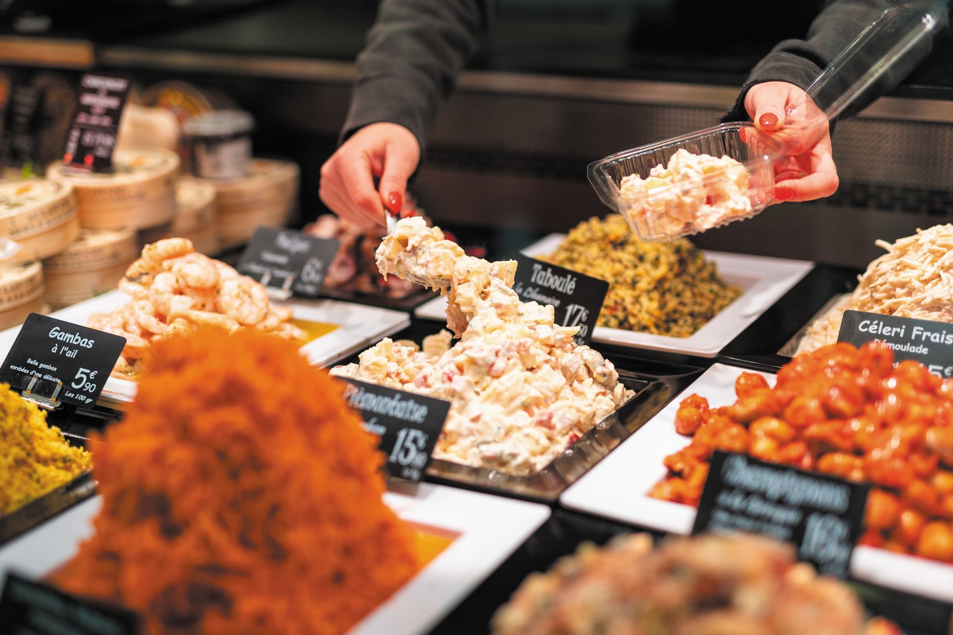 Comptoir de boulangerie avec assortiment de salades colorées et service à la main de portions servies dans des plateaux blancs.