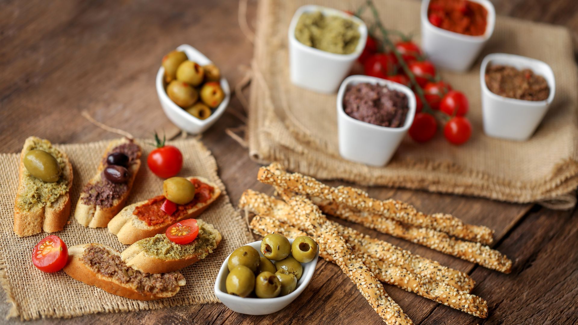 Assortiment de bruschettas et de gressins avec olives, tomates cerises et petits ramequins de condiments sur une table rustique.