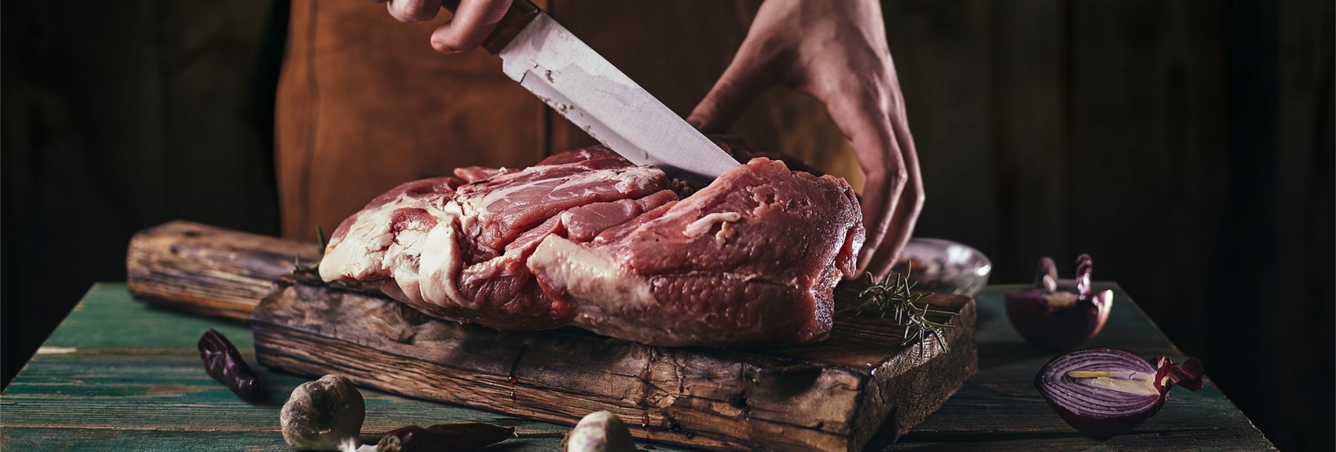 Des mains découpent de la viande crue sur une planche à découper en bois avec un couteau, une nappe verte en dessous