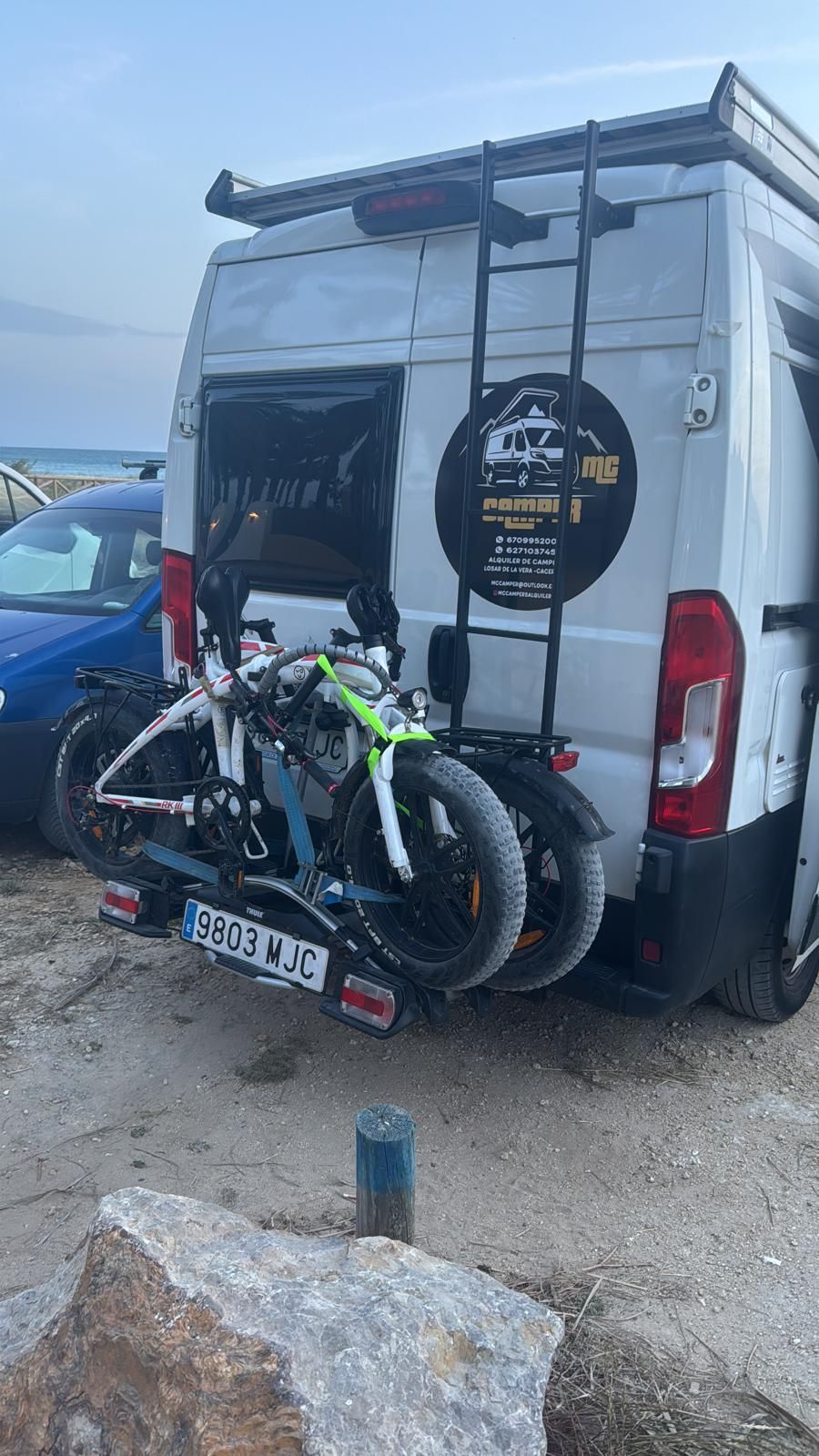 Equipamiento para bicicletas sujeto a la parte trasera de una furgoneta camper blanca en una zona de aparcamiento rocosa al aire libre.