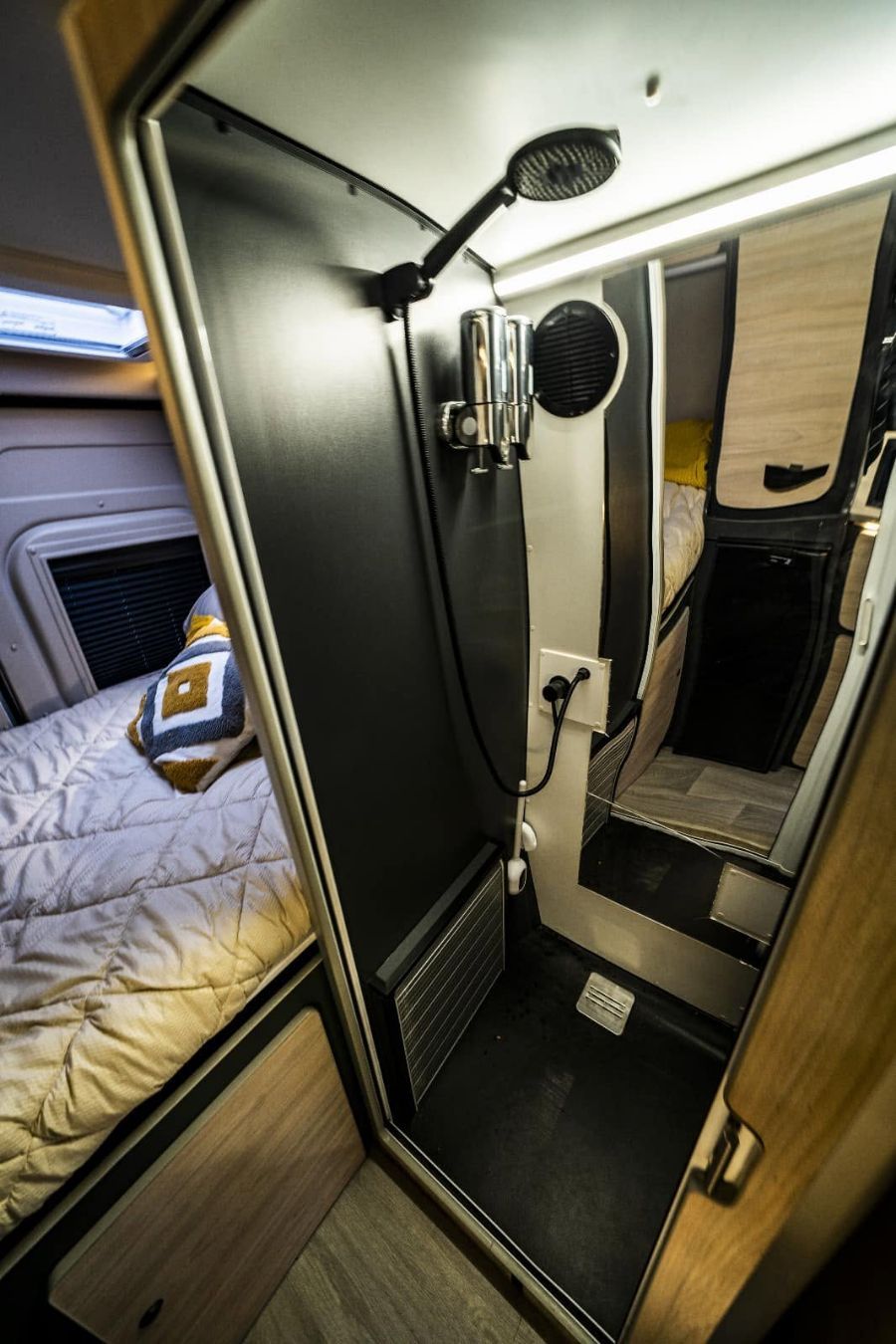 Baño compacto para autocaravana con cabina de ducha negra, cabezal de ducha de mano y armario con espejo; la cama es visible al lado.