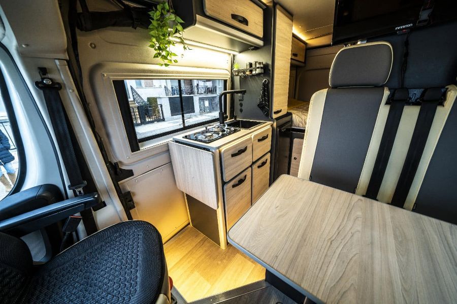 Interior de furgoneta camper compacta con cocina americana, mesa de madera y asientos en blanco y negro.