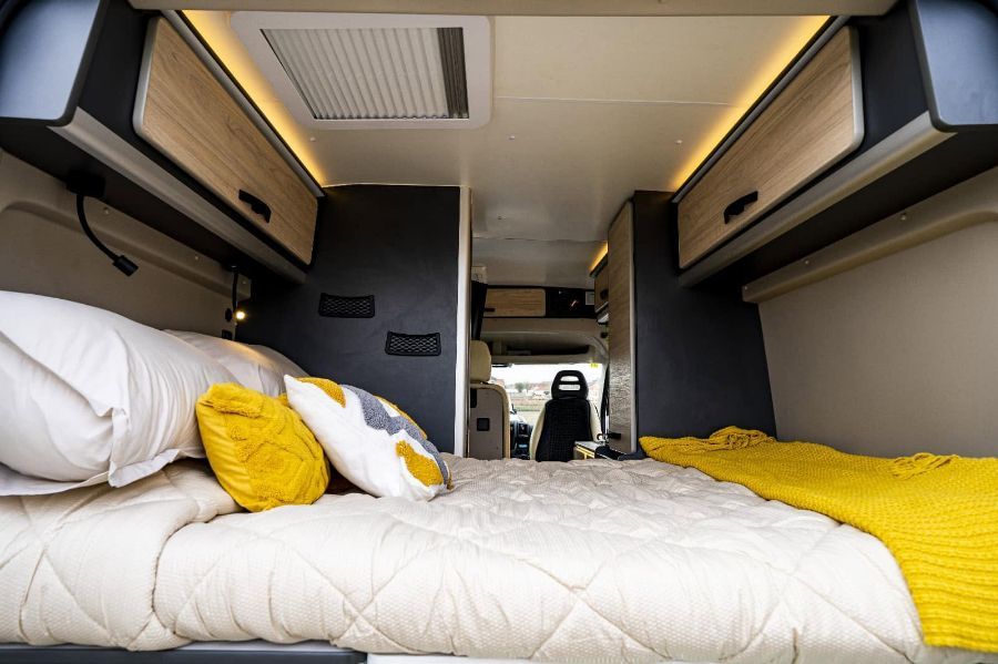 Interior de una furgoneta camper con cama, ropa de cama blanca, almohadas amarillas y armarios superiores.