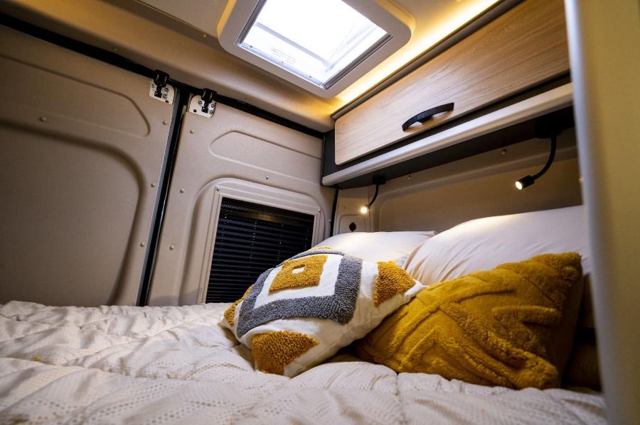Acogedor rincón para dormir en una caravana, con una cama blanca, almohadas amarillas y espacio de almacenamiento superior bajo una claraboya.