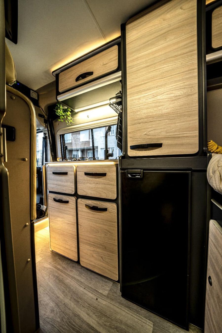 Cocina compacta para autocaravana con armarios de madera clara, nevera negra e iluminación LED cálida.