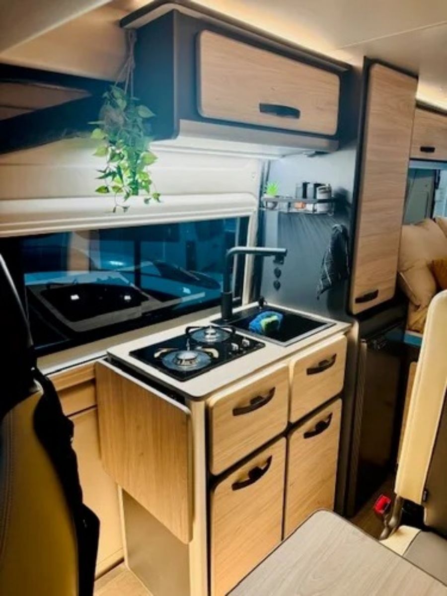 Cocina compacta para caravana con armarios de madera, placa de cocina, fregadero, nevera y una pequeña ventana con una planta colgante.