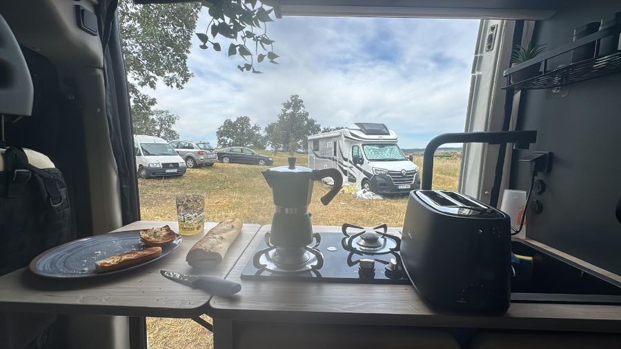 Cafetera preparada sobre una mesa junto a la puerta abierta de una autocaravana, con hervidor, hornilla, taza y plato, con vistas a una zona de aparcamiento cubierta de césped.