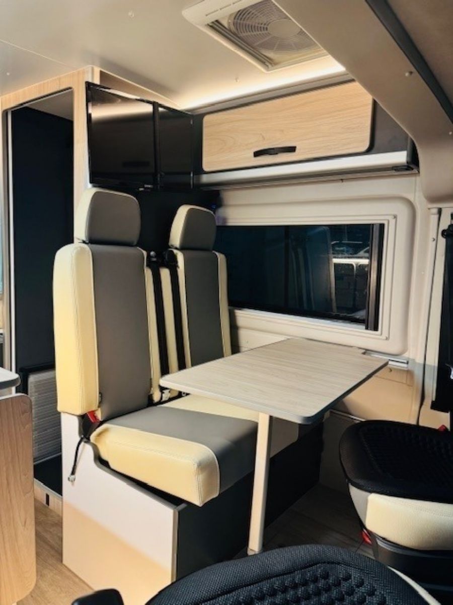 Interior de furgoneta camper compacta con asientos grises, mesa plegable, espacio de almacenamiento superior y zona de estar junto a la ventana.