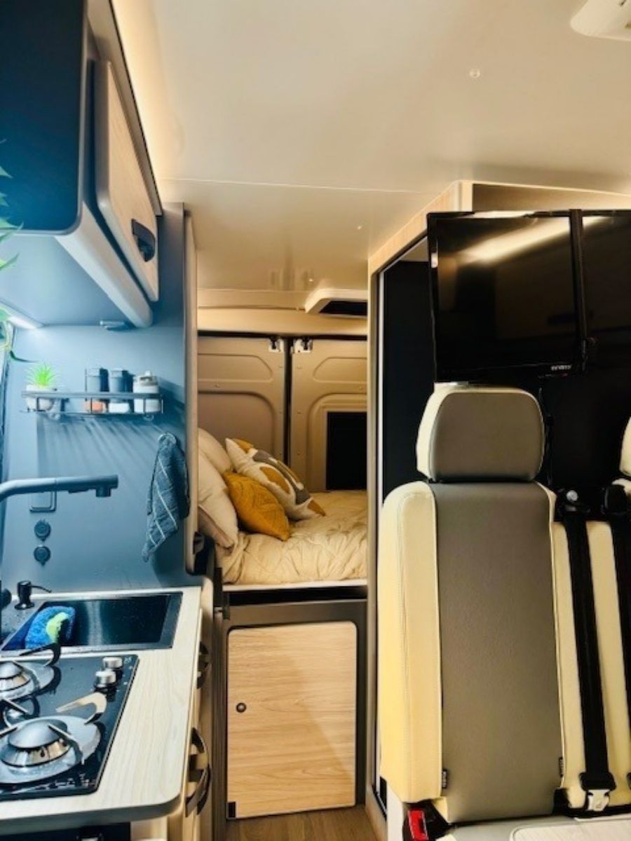 Interior de furgoneta camper compacta con cocina americana, sofá cama y espacio de almacenamiento superior en una combinación de colores beige y negro.