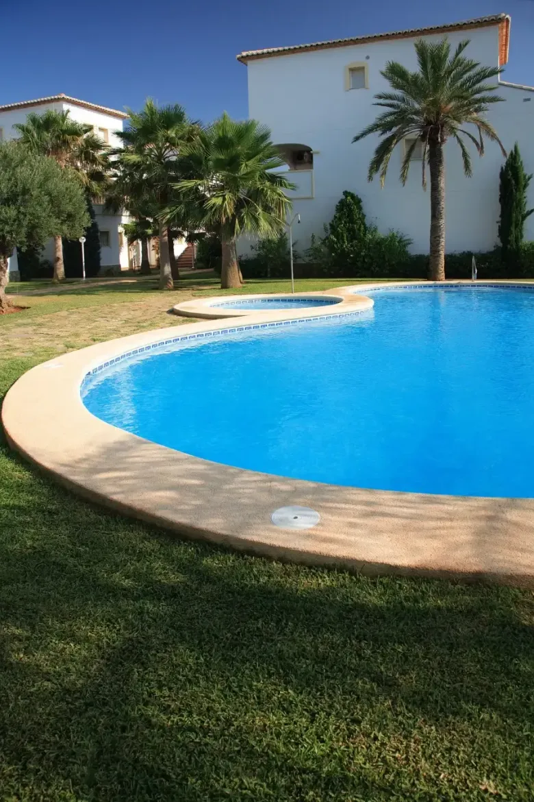 Piscina azul rodeada de césped verde, cerca de edificios blancos y palmeras. Día soleado.