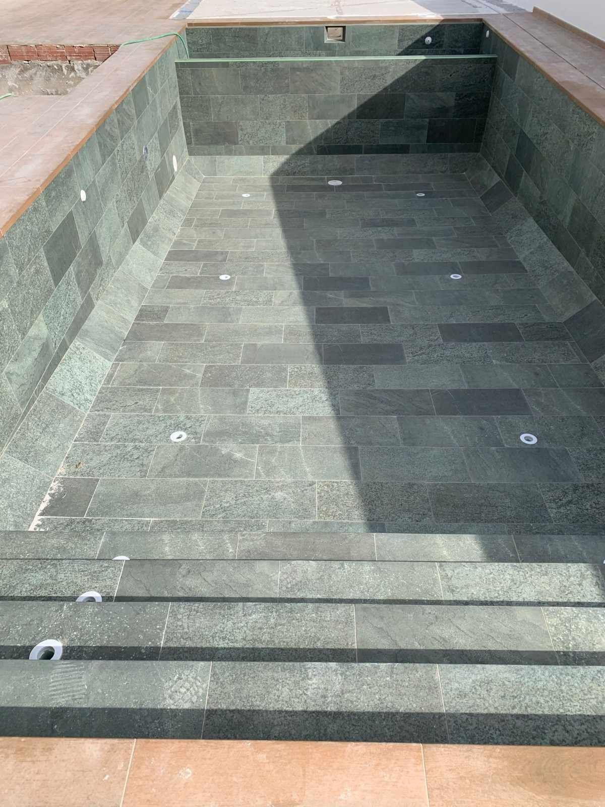 Piscina rectangular vacía con azulejos de color verde oscuro, escalones y accesorios de iluminación.