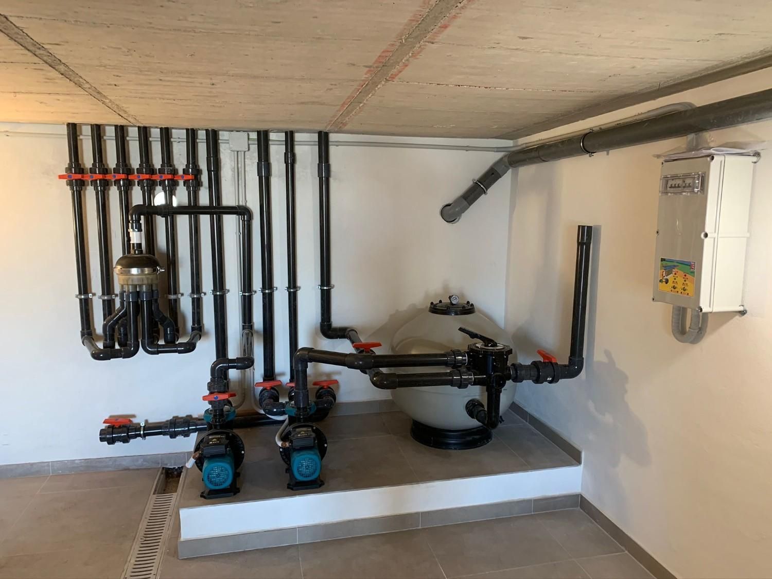 Sistema de filtración de piscina con tuberías negras, válvulas rojas y bombas en un sótano.