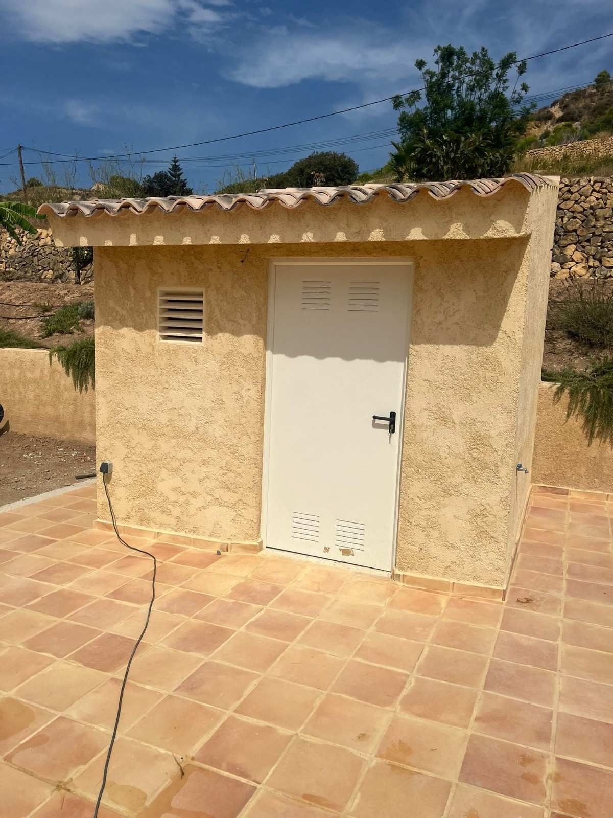 Pequeño edificio de estuco beige con puerta blanca, patio de baldosas y fondo de montaña.
