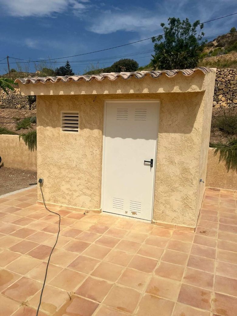 Pequeño edificio de estuco beige con puerta blanca, patio de baldosas y fondo de montaña.