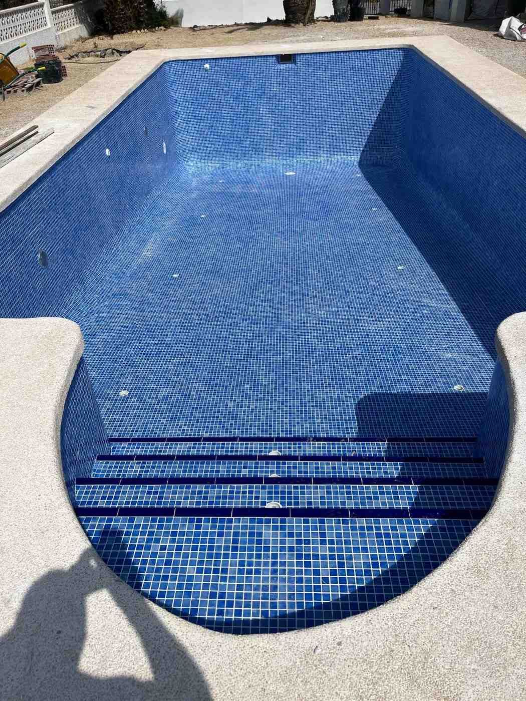 Piscina vacía con interior de azulejos azules y escalones. Borde de hormigón y luz solar intensa.