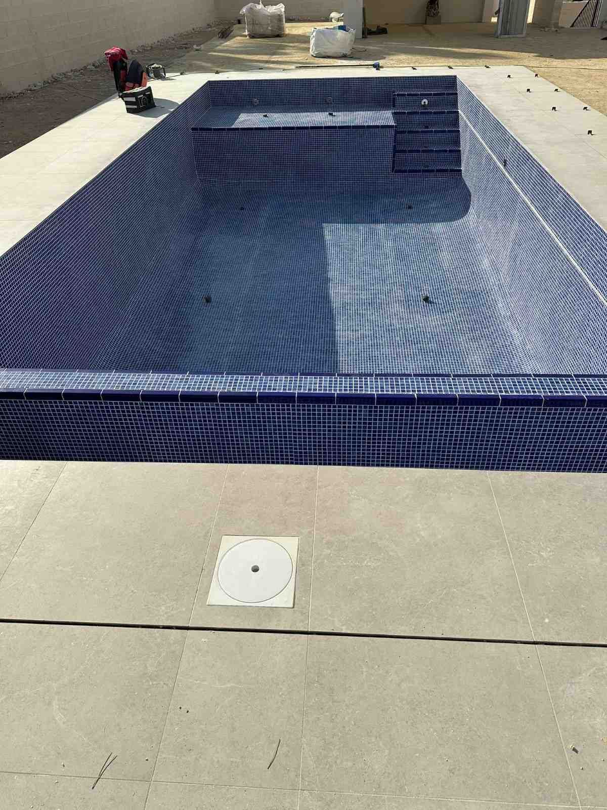 Piscina rectangular vacía con mosaicos azules, colocada en hormigón, con un área de patio de color claro a su alrededor.