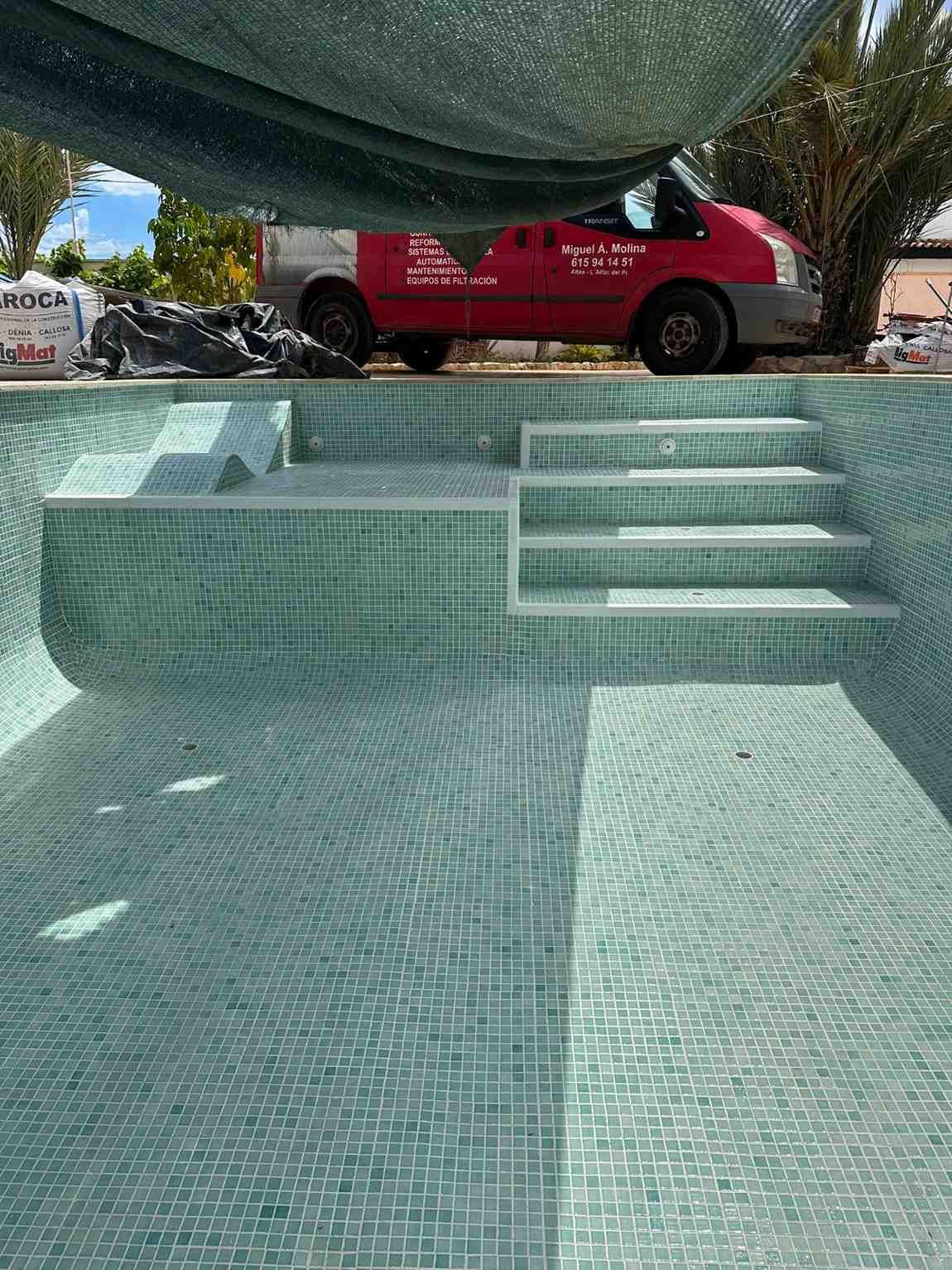 Piscina vacía con azulejos turquesas y escalones incorporados; camioneta roja estacionada cerca.