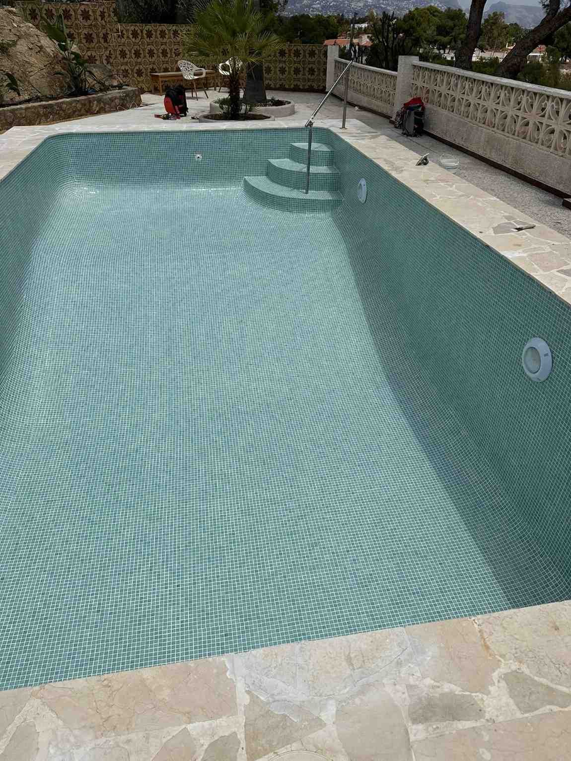 Piscina llena de agua, revestida con mosaicos verdes, escalones en un extremo, patio de piedra alrededor.
