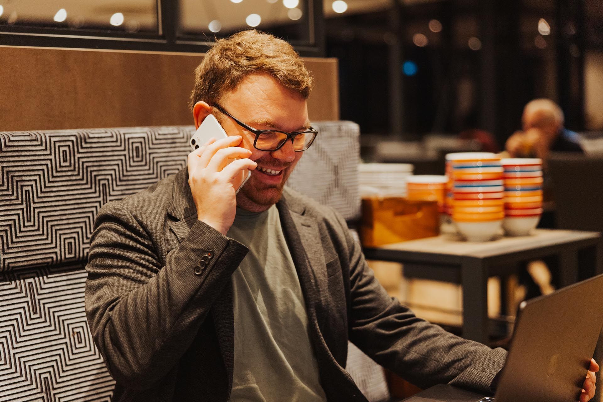 Mann telefoniert, lächelt und benutzt seinen Laptop in einem Café mit bunten Kaffeetassen im Hintergrund.