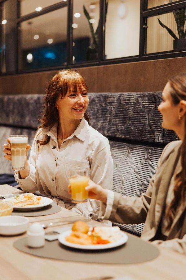 Zwei Personen stoßen an einem Restauranttisch an, neben dem Teller mit Essen und Kaffeetassen stehen.