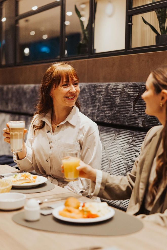 Zwei Personen stoßen an einem Restauranttisch an, neben dem Teller mit Essen und Kaffeetassen stehen.