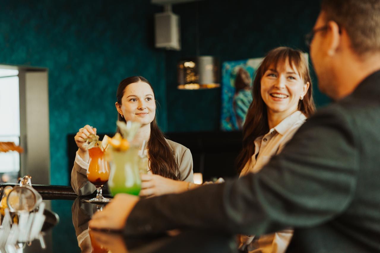 Drei Personen sitzen an einer Bar, vor sich stehen bunte Cocktails, und sie lächeln, während sie sich angeregt unterhalten.