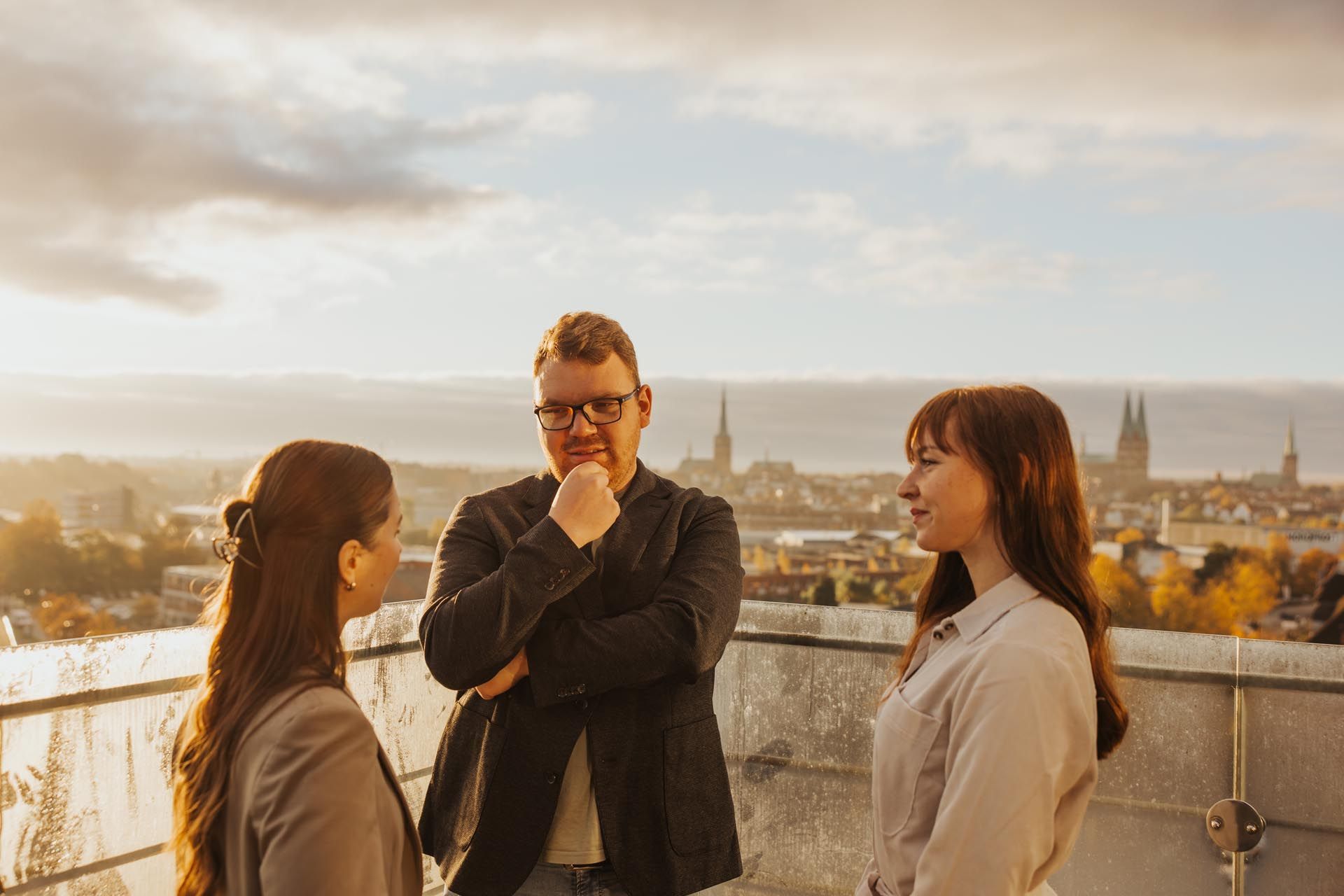 Drei Personen unterhalten sich auf einem Dach mit Blick auf die Stadt bei Sonnenuntergang.