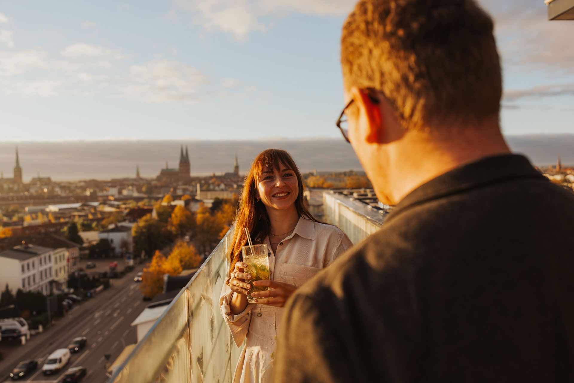 Ein Paar auf einer Dachterrasse bei Sonnenuntergang, lächelnd über die Skyline der Stadt mit einem Glas Wein.