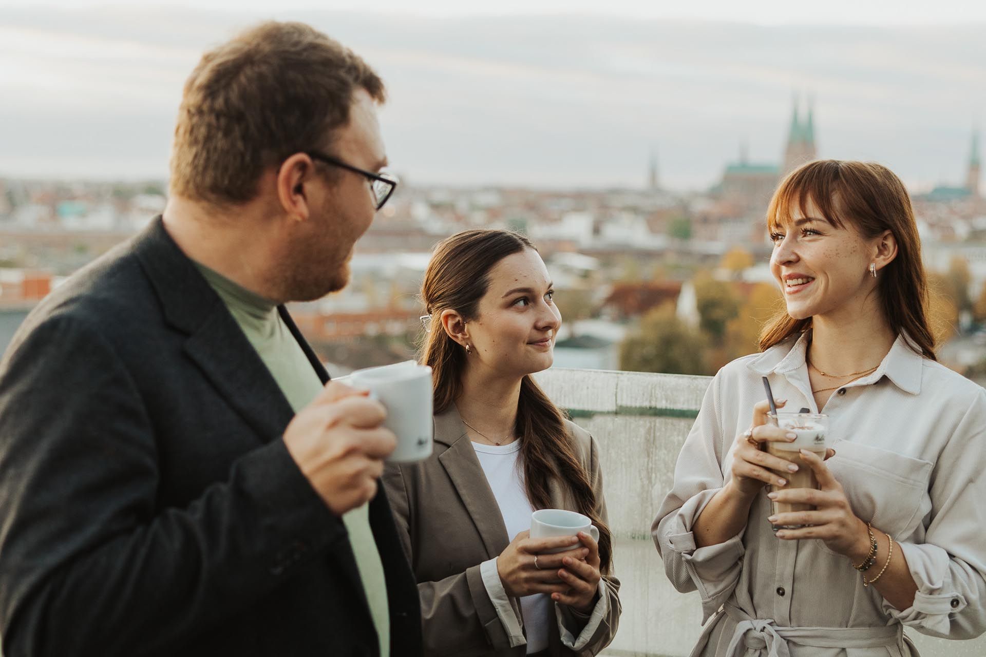 Drei Personen unterhalten sich auf einem Balkon mit Kaffeetassen und blicken auf die Skyline der Stadt.